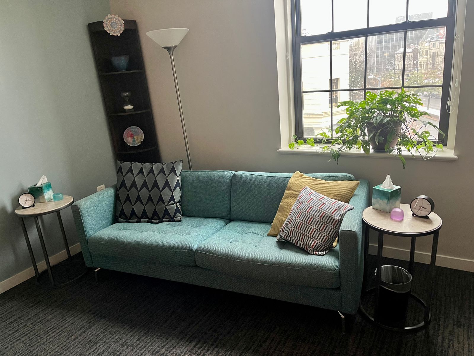 Therapy office with a teal sofa, patterned pillows, two side tables, and a window with a leafy plant